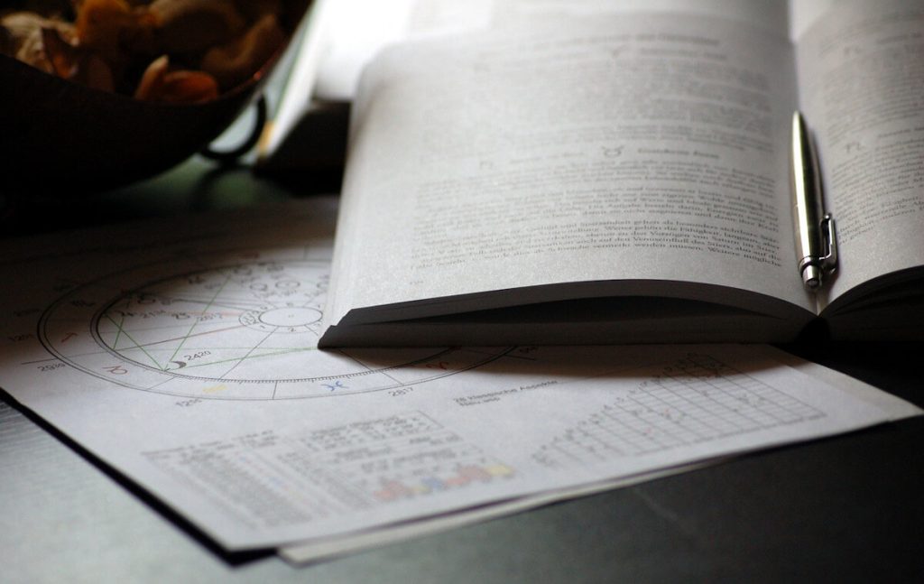 Open astrology book with a pen, resting on printed birth charts and aspect tables