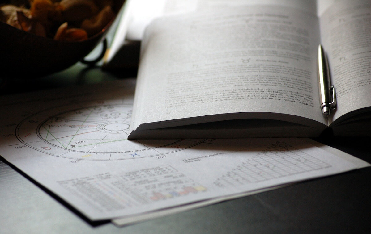 Open astrology book with a pen, resting on printed birth charts and aspect tables