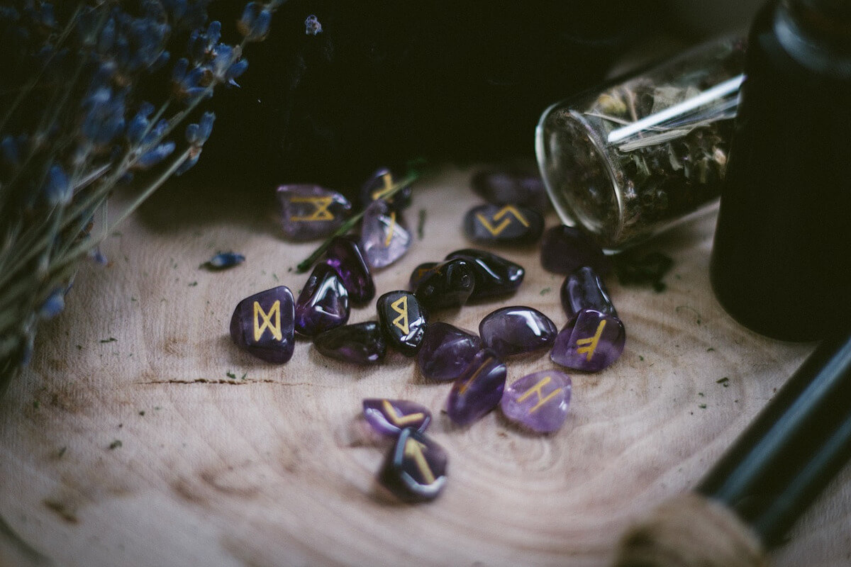 Purple stone runes engraved with gold symbols on a slice of wood, with lavender and a jar of dried herbs