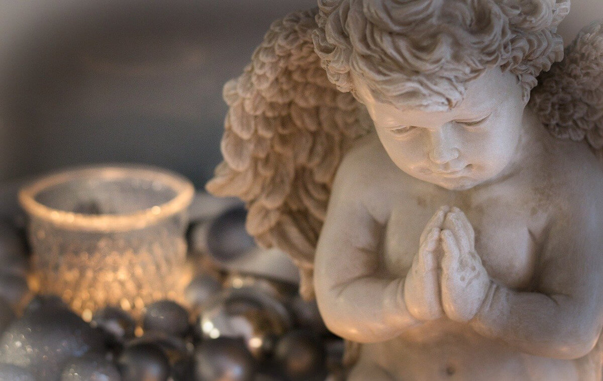 Stone cherub figurine with hands joined in prayer, beside a candle in a glass holder