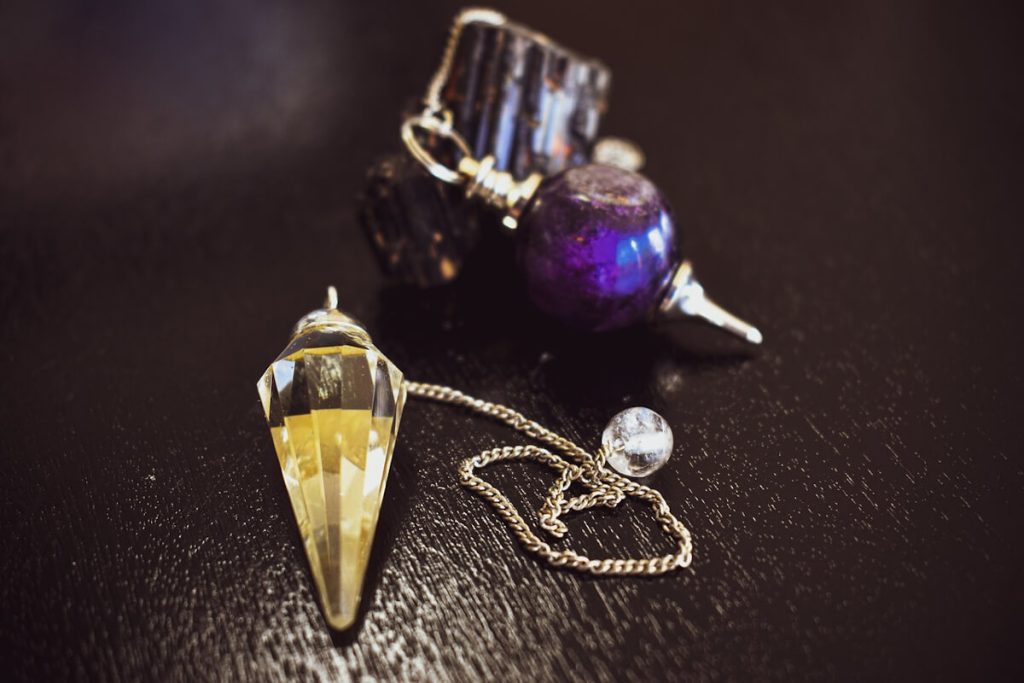 Péndulo de cristal facetado con su cadena junto a un péndulo de esfera violeta y piedras oscuras en bruto