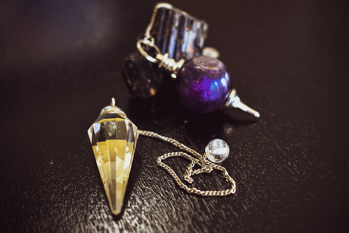 Péndulo de cristal facetado con su cadena junto a un péndulo de esfera violeta y piedras oscuras en bruto
