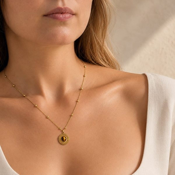 Pendentif médaille en œil de tigre porté à mi-poitrine, sur une chaîne fine à petites boules.